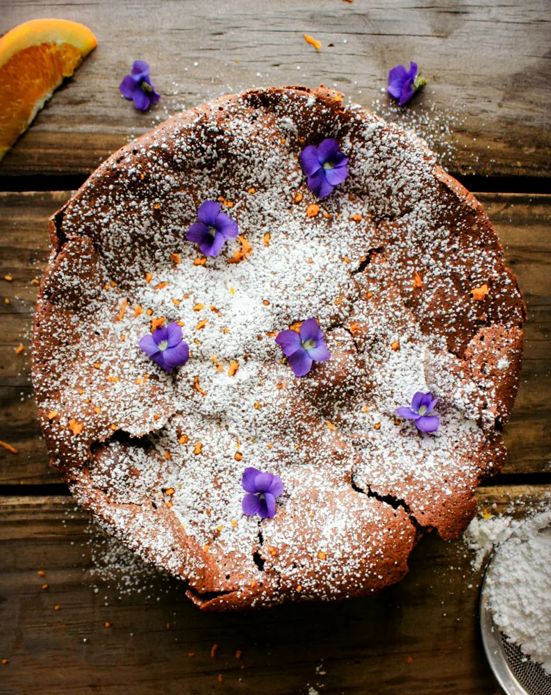 Flourless Chocolate Orange Torte. Covered in powder sugar and decorated with orange shavings and purple flowers.