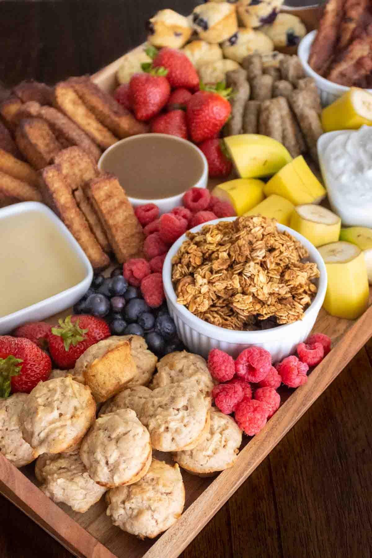 Breakfast charcuterie board with mini muffins, bacon, french toast sticks, granola, and fruit.