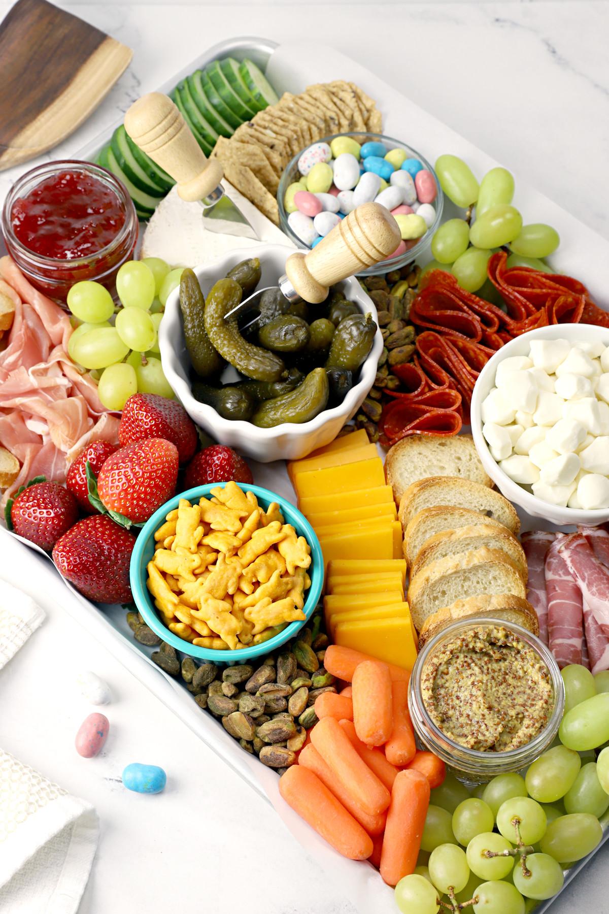 Easter charcuterie board with pickles, grapes, strawberries, meats, crackers, carrots, breads, and jam.