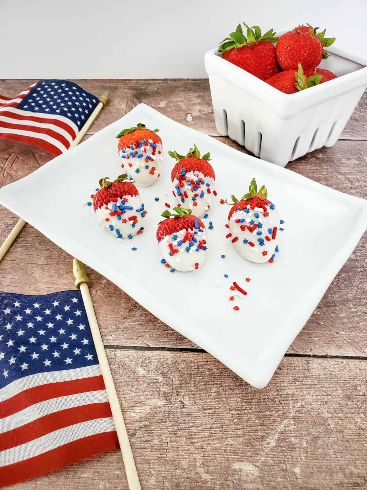 White chocolate dipped strawberries covered in sprinkles on a white rectangular tray. There's a white bowl of strawberries and small American flags.