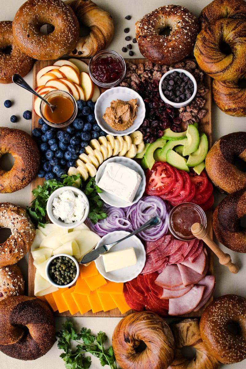 Bagel brunch board with different types of bagels, avocado, bananas, blueberries, chocolate, raisins, tomatoes, onions, cheeses, meats, and honey.