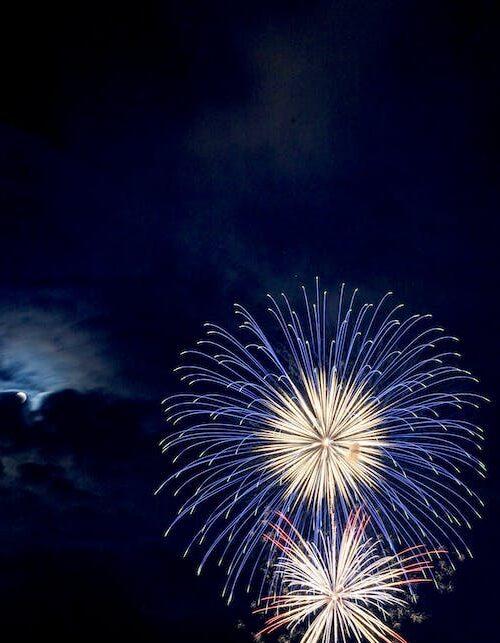Fireworks in the night sky with the moon peaking out behind a cloud