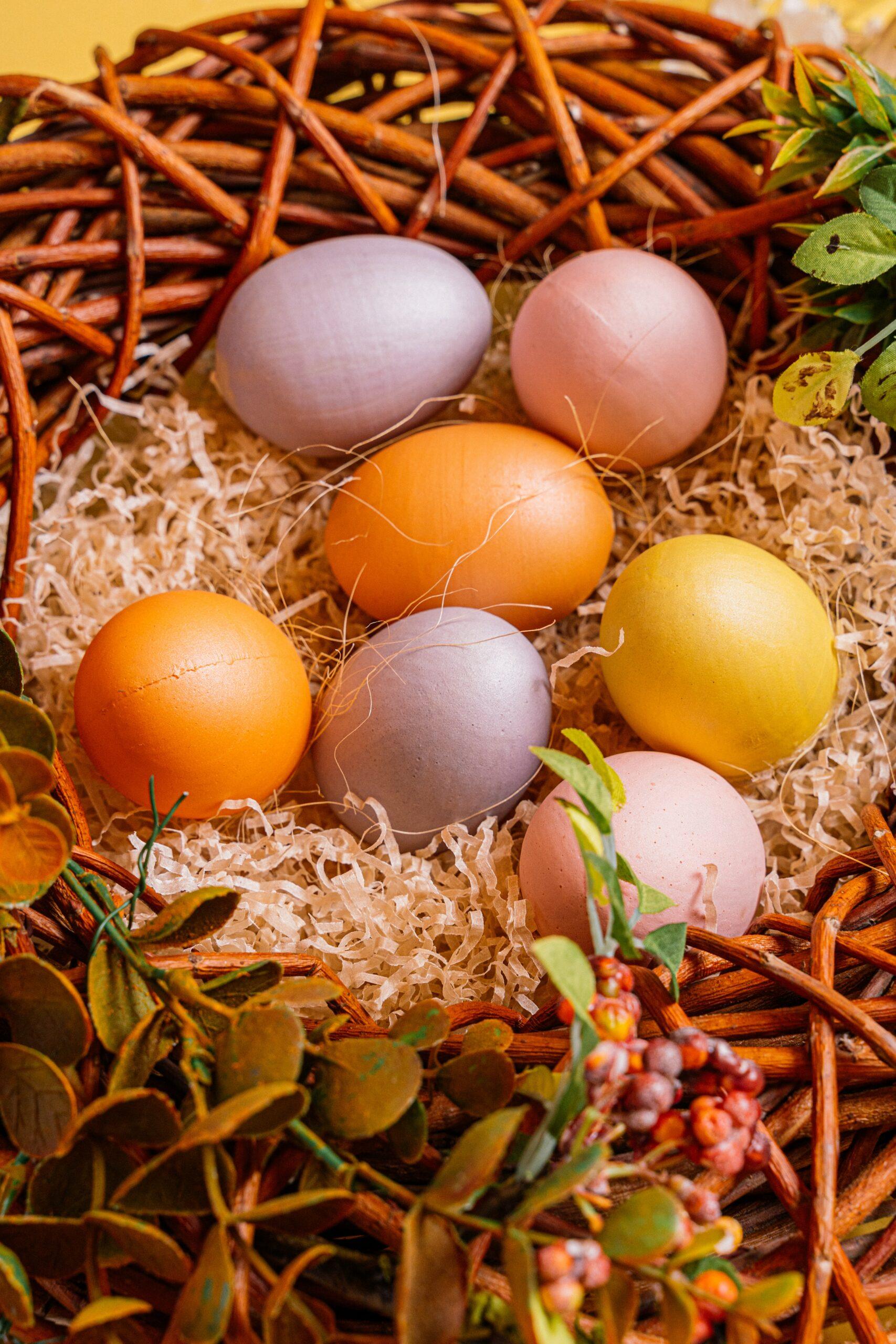 Pastel orange, purple, pink, and yellow dyed eggs in a wicker basket with green foliage and fake grass