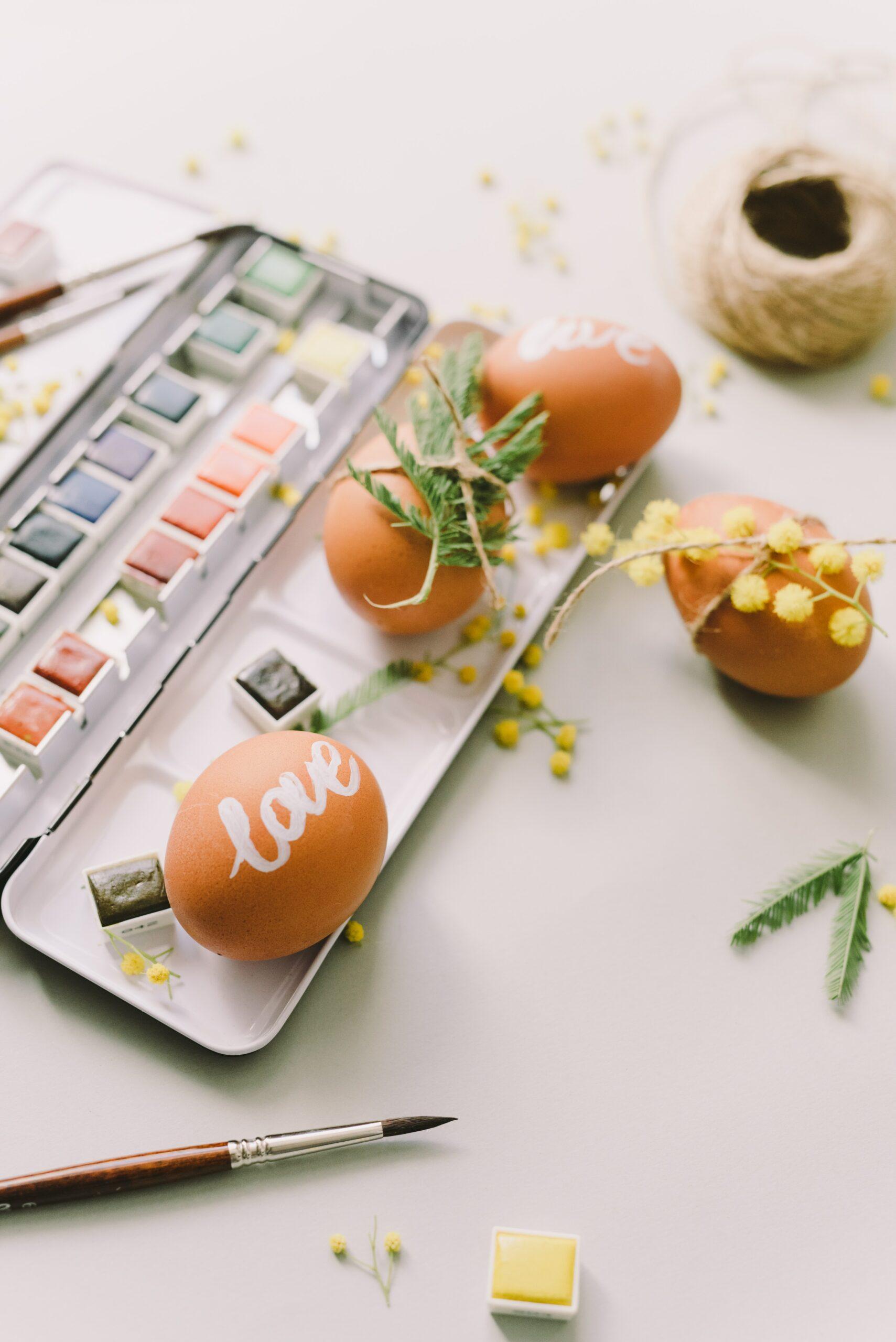 Brown eggs painted with the word "love" in white paint. The eggs are sitting on a paint pallet with some flowers and greenery.