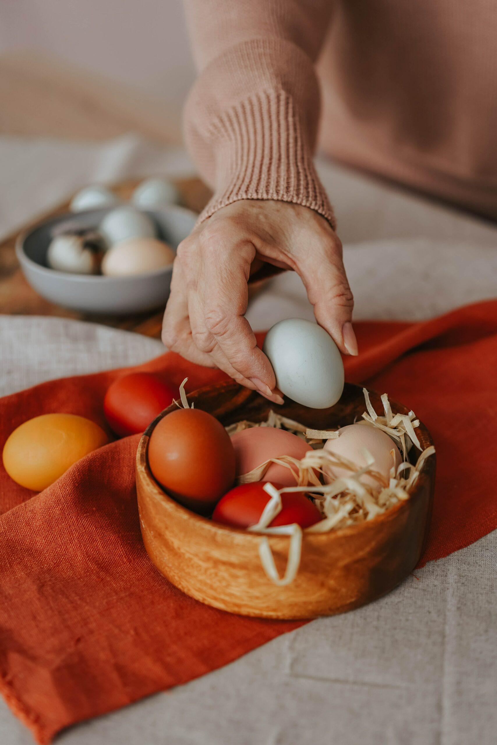 A hand placing a light blue egg into a wooden bowl with other eggs (pink, red, and orange)