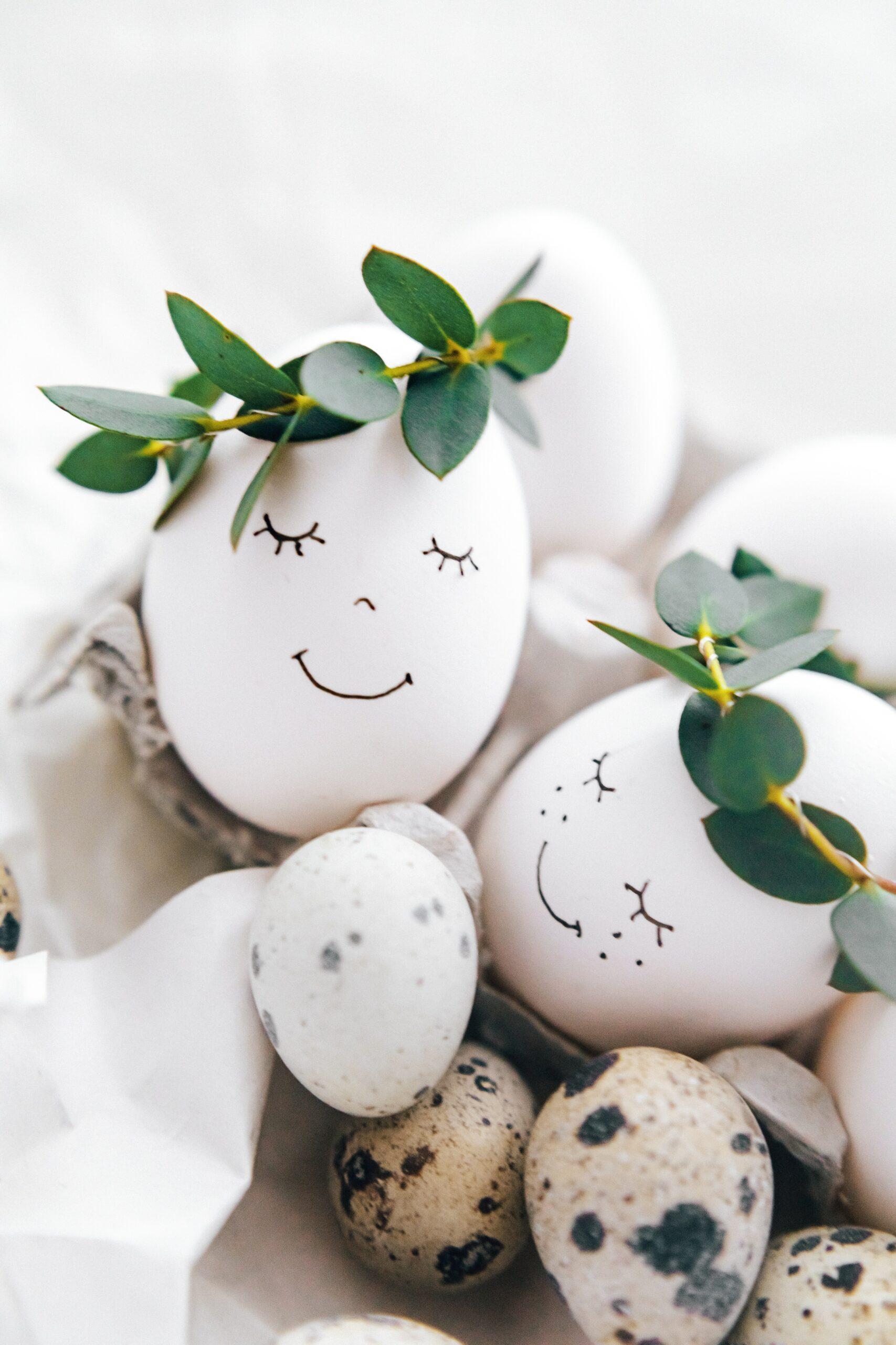 White eggs in a carton with drawn-on faces with a black marker. The eggs have greenery crowns