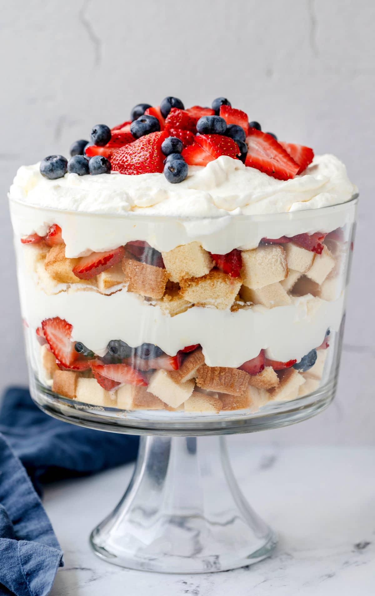 Red white and blue trifle in a beautiful glass bowl so you can see each colorful layer. It's topped with whipped cream, strawberries, and blueberries