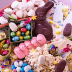 Top down view image of an easter charcuterie board filled with chocolate bunny, peeps, popcorn, cadbury eggs, and easter chocolates - square image