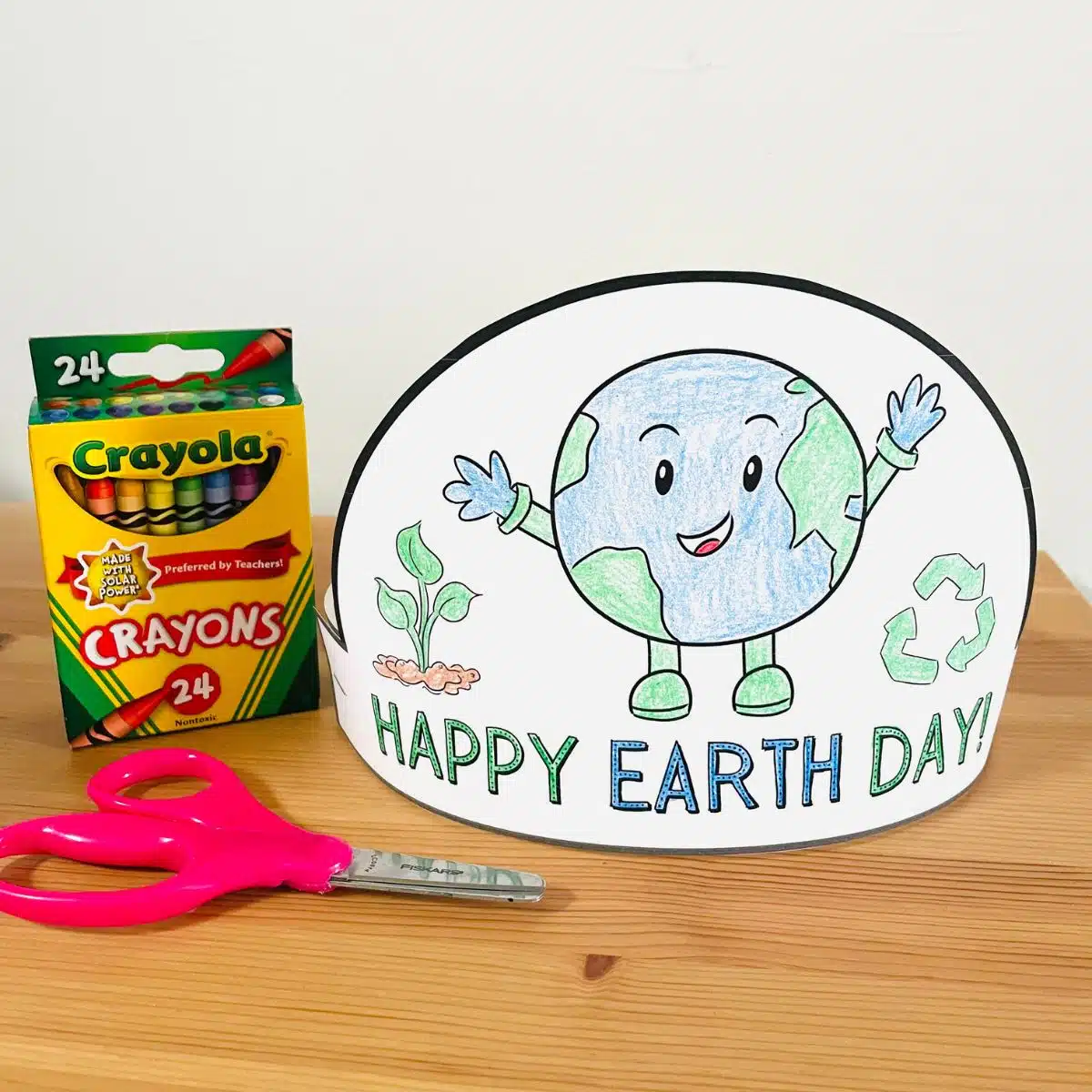 An earth day hat colored with green and blue next to a box of crayons and scissors