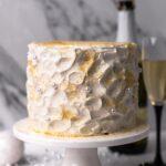 Champagne Cake on a white cake stand with a champagne bottle in the back
