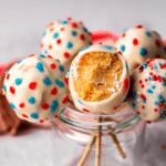 Close-up of festive cake pops decorated with red, white, and blue sprinkles, with one pop showing a bite revealing a soft, golden interior, displayed in a glass jar for a patriotic celebration