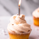 square image of champagne cupcake with champagne frosting with candle sticking out of the top. Side view to show the silky texture of the frosting.