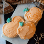 Halloween sugar cookies with orange cream cheese frosting and green frosting stems staggered on top of each other with fall decorations in the background